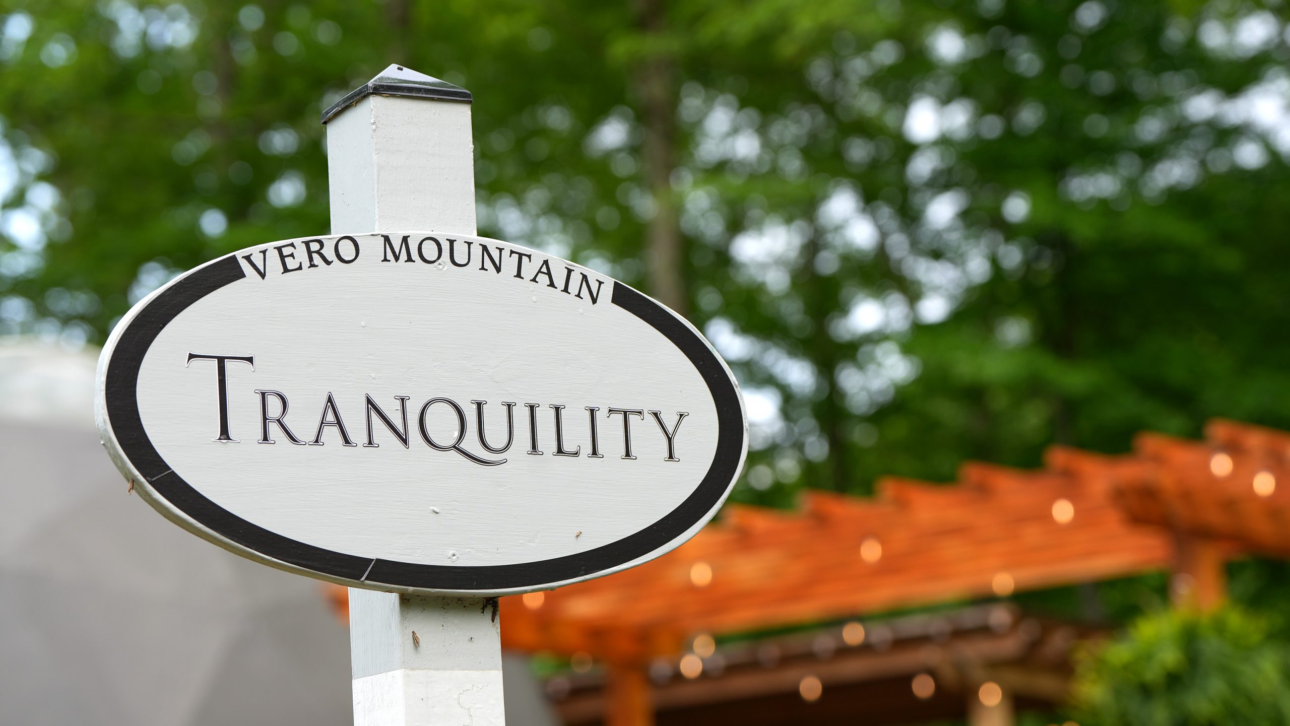 A white oval sign reading Vero Mountain Tranquility stands in front of green trees and a wooden pergola with string lights, offering an inviting backdrop perfect for photography & videography services.