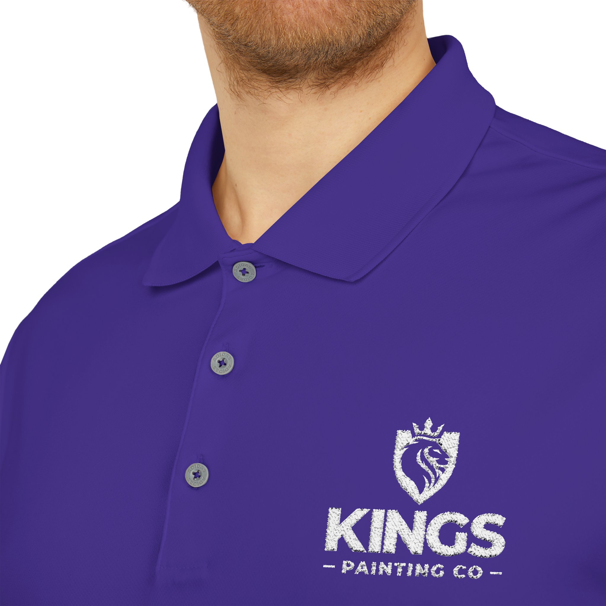 A person wearing the Kings Painting Co. Performance Polo by Under Armour®, featuring a white embroidered crowned lion head and text logo on the chest. Only the lower face and upper torso are visible.