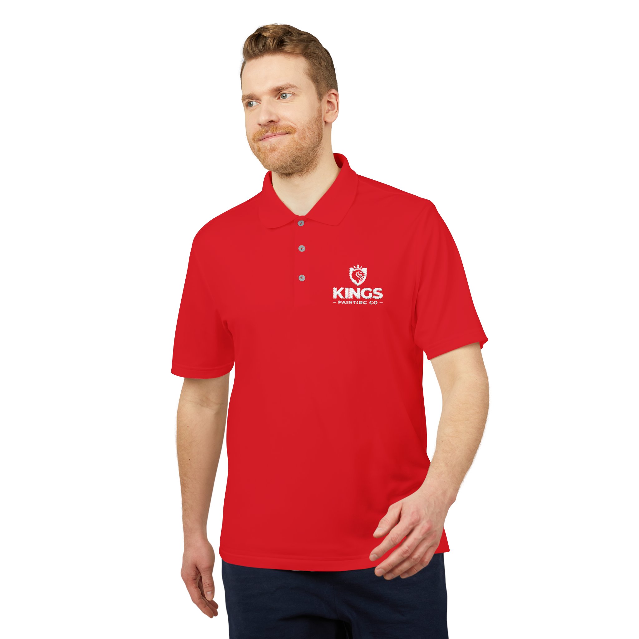 A man with light skin and brown hair is wearing the Kings Painting Co. Performance Polo by Under Armour®, featuring a white KINGS and crown logo on the left chest. He stands facing slightly to the left, smiling, with his right hand at his side and his left arm bent.